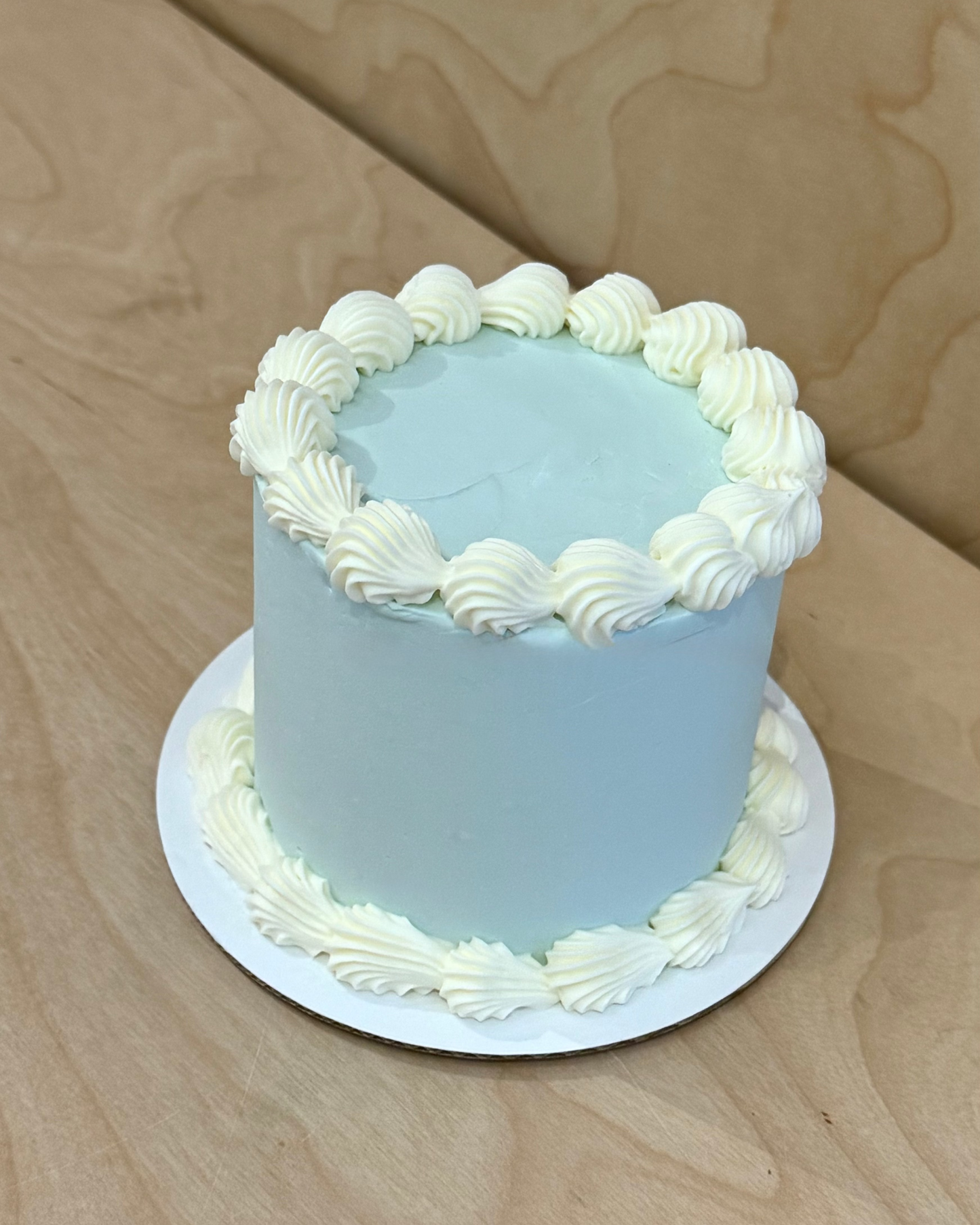 A small round cake with smooth light blue frosting and piped white frosting borders on top and around the base, sitting on a white round board over a light wood surface.